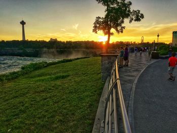 People on footpath by road against sky during sunset