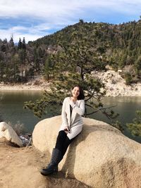 Full length portrait of young woman sitting by lake on rock