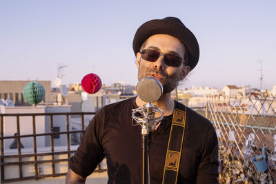 Portrait of young man wearing sunglasses singing by microphone standing against sky