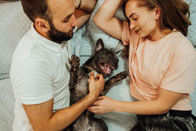 Happy couple man and woman laying on the bed with their dog, funny french bulldog having fun