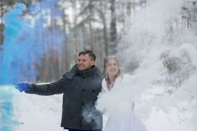 Smiling wedding couple holding distress flares while standing at forest during winter