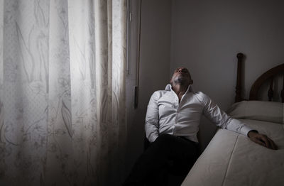 Adult man in white shirt in room sitting on chair beside bed