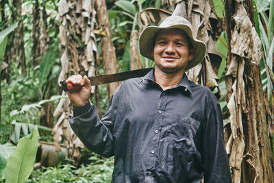 Positive hispanic man in shirt and bucket hat touching shoulder with machete and looking at camera with smile during work on ecological banana farm in costa rica