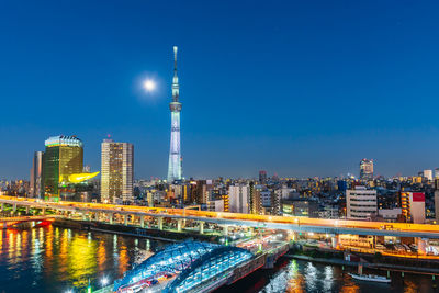 Illuminated bridge over river by buildings against sky at night