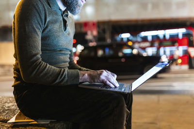 Freelancer using laptop sitting on bench in city at night