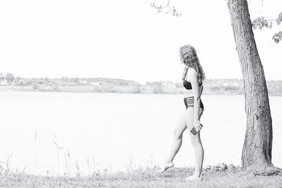 Side view of woman wearing swimwear while standing at lakeshore against clear sky