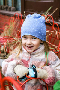 A young girl with blonde hair smiles in a blue hat with white polka dots, a colorful jacket. 