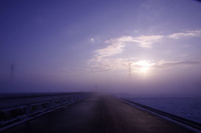 Empty road leading towards river in foggy weather at sunset