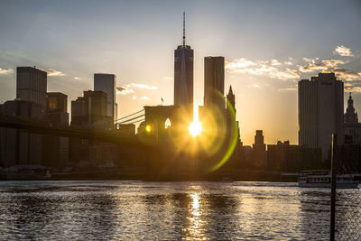 Modern buildings by river against sky during sunset