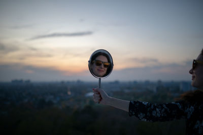 Side view of woman holding mirror while standing against sky during sunset