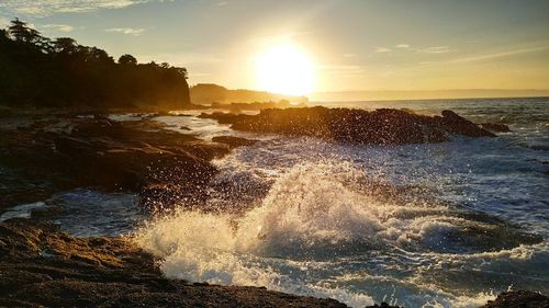 Sea waves splashing on rocks against sky during sunset