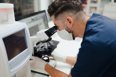 Side view of crop anonymous male medic in uniform and mask using microscope while working in lab