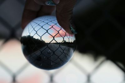 Cropped hand of woman holding crystal ball with fence and road reflected on it during sunset