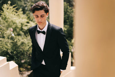 A beautiful young man, the groom in an elegant wedding suit, stands posing in the city's old park