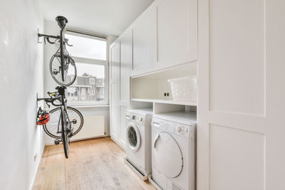 Modern white electric washing and dryer machines placed near white cabinets and basket in light laundry room with window and bicycle