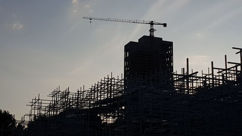 Low angle view of silhouette crane by building against sky