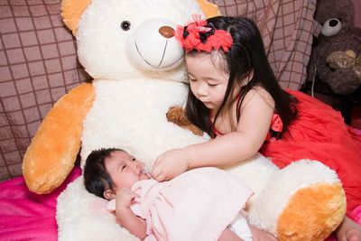 High angle view of sister putting pacifier in baby girl mouth while lying on teddy bear at bedroom