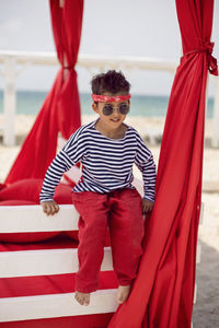 Stylish baby boy in a striped t-shirt and sunglasses stands at the red beach sunbed