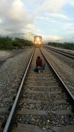 Rear view of woman sitting on railroad track by illuminated train against cloudy sky