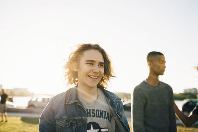 Smiling young man looking away while friend standing in background against clear sky during sunny day