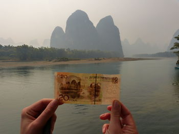 Close-up of hands holding paper currency by river against mountains