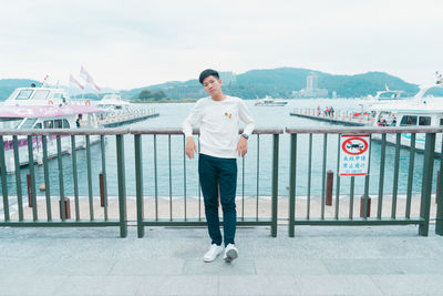 Portrait of young man standing on railing against sky