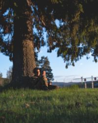 Man sitting on field by trees against sky
