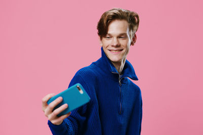 Portrait of smiling young man using mobile phone while standing against pink background