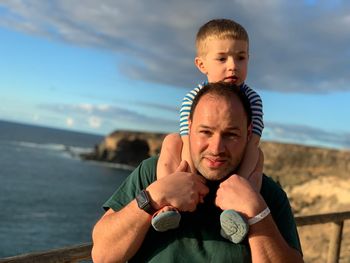 Portrait of father carrying son on shoulders against sea and sky