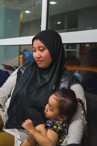 Mother wearing hijab with daughter sitting on chair at airport