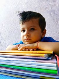 Boy with stacked books looking away while sitting against wall at home