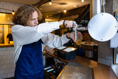 Concentrated young male server in apron filling glass with fresh cold beer from tap while standing at bar counter