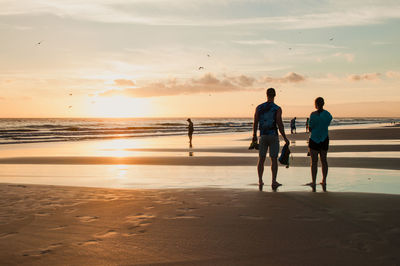 Rear view of men walking on beach against sky during sunset