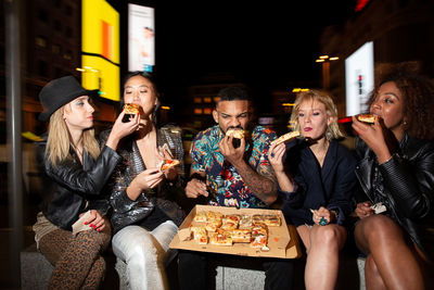 Young multiracial partners with crossed legs enjoying tasty baked food from takeaway box during celebration in town in evening