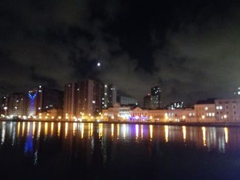 Illuminated buildings by river against sky at night