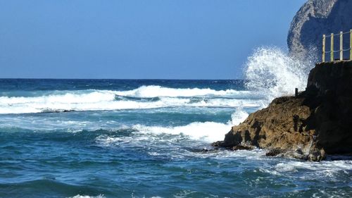 Waves splashing on rocks by sea against clear sky