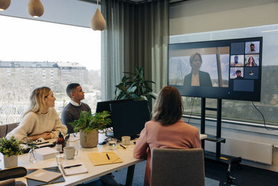 Male and female colleagues doing video call with businesswoman over tv in coworking office