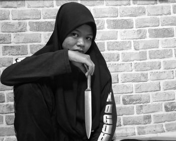 Portrait of young woman holding knife against brick wall