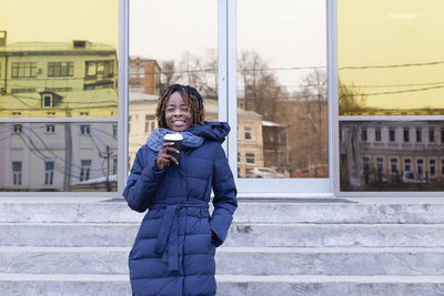 Portrait of woman holding coffee cup standing by entrance of building