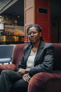 Portrait of confident young female entrepreneur wearing eyeglasses sitting on chair in hotel lounge