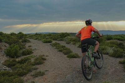 Rear view of man cycling on dirt road against cloudy sky during sunset