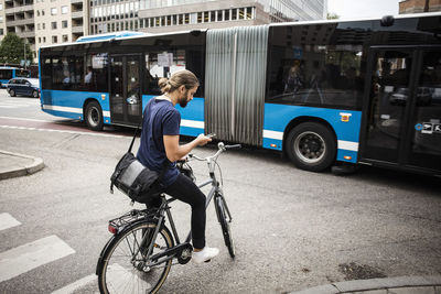 Man with bicycle using mobile phone while standing on city street against articulated bus
