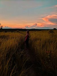 Side view of woman amidst plants on field against sky during sunset