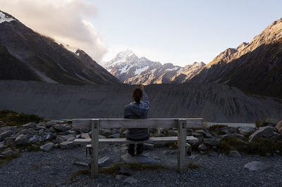 Rear view of woman photographing mountains while sitting on bench against cloudy sky