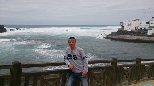 Portrait of man leaning on railing by sea against sky