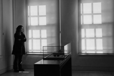 Full length of thoughtful woman standing in living room by sunlight on wall