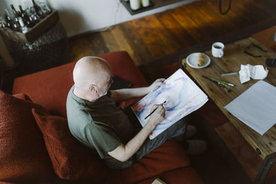 High angle view of senior man painting face over paper while sitting on sofa at home