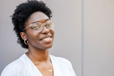 Delighted african american female in white wear and glasses and work uniform looking at camera with smile while standing on street near wall