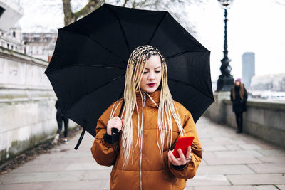 Woman with umbrella using mobile phone while walking on footpath