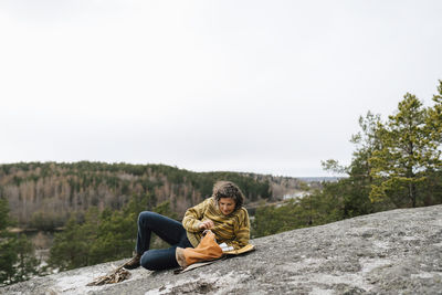 Young woman sitting on rock against sky